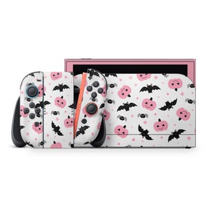 Può includere: Una console Nintendo Switch bianca e i suoi controller con un motivo a tema Halloween. Il design presenta zucche rosa, pipistrelli neri e stelle su sfondo bianco. La console ha una cornice rosa.