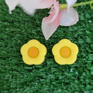 May include: Two yellow flower-shaped earrings with a smaller yellow circle in the center. The earrings are on a green surface.