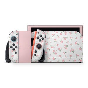 May include: A pink and white Nintendo Switch console with matching Joy-Con controllers. The console and controllers are decorated with a floral pattern of pink flowers and green leaves on a white background. The console is shown on a white background.