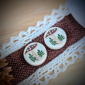 May include: Two round, cream-colored mushroom earrings with brown caps and white polka dots. The earrings have a light beige stem and green leaves. They are displayed on a brown ribbon with white lace trim.