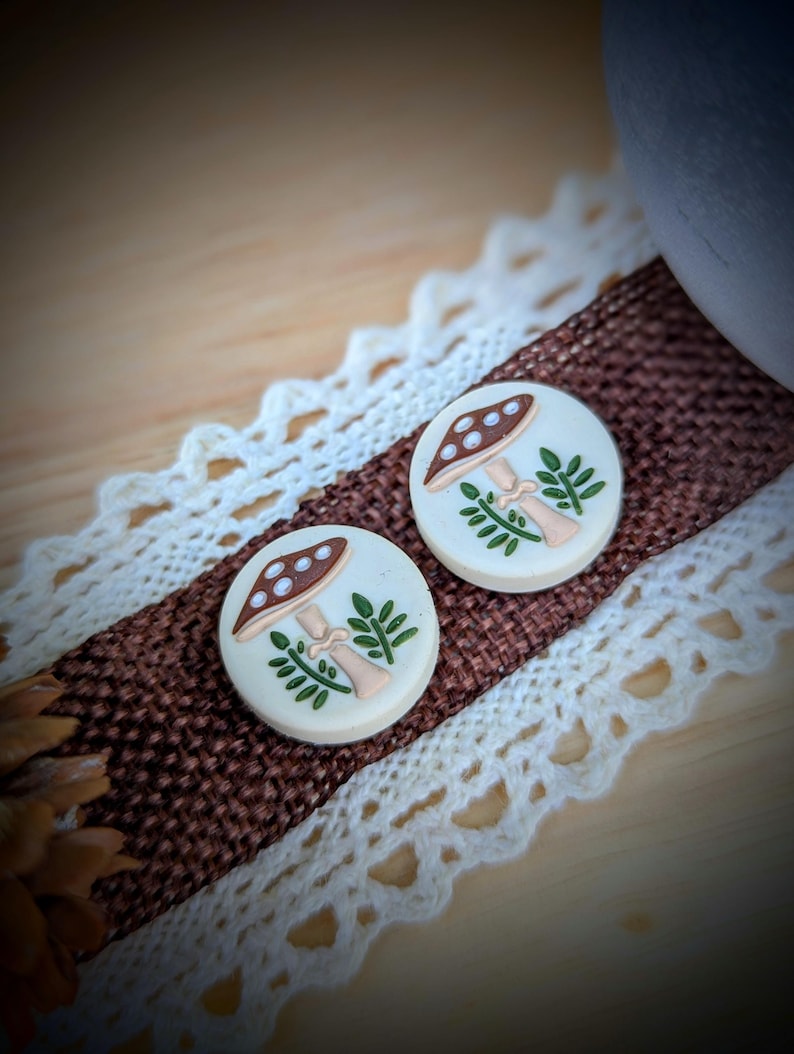 May include: Two round, cream-colored mushroom earrings with brown caps and white polka dots. The earrings have a light beige stem and green leaves. They are displayed on a brown ribbon with white lace trim.