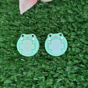 May include: Two green frog-shaped silicone buttons with white eyes and a pink mouth. The buttons are on a green artificial grass background.