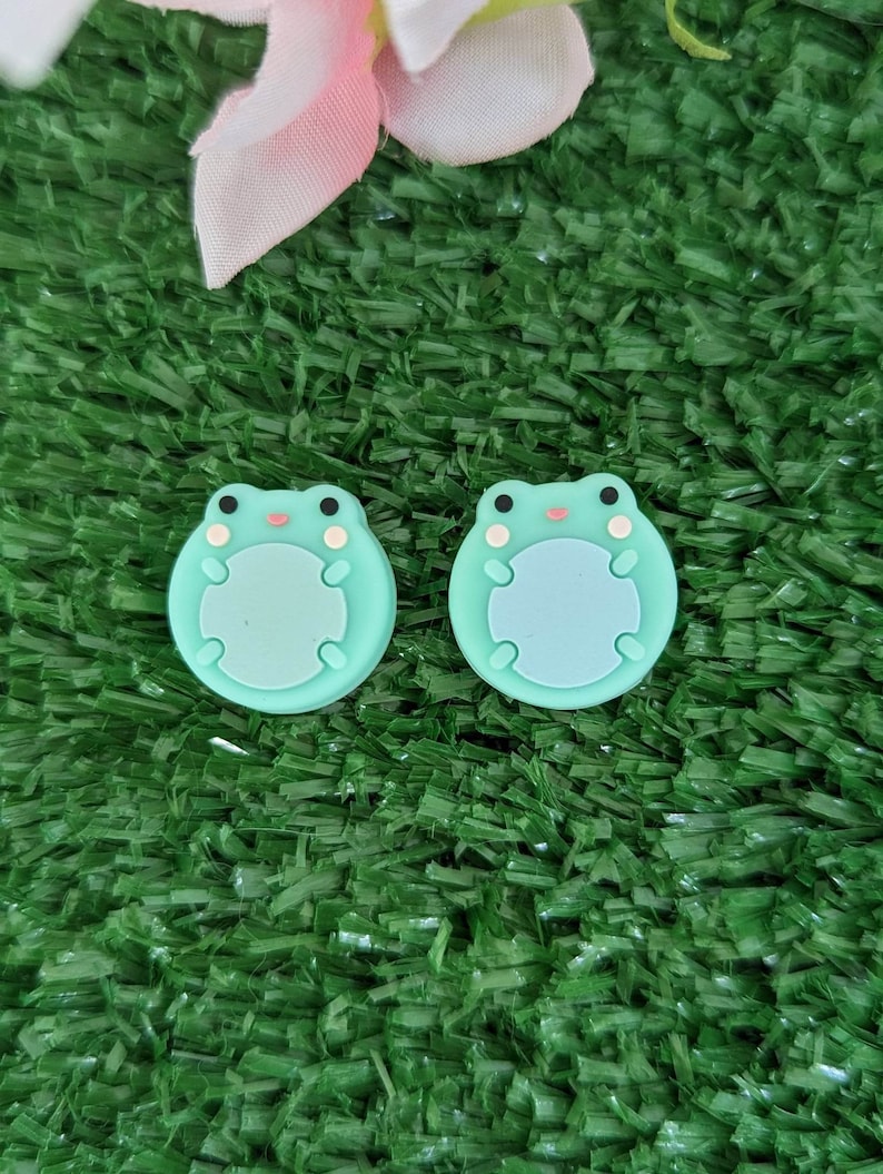 May include: Two green frog-shaped silicone buttons with white eyes and a pink mouth. The buttons are on a green artificial grass background.