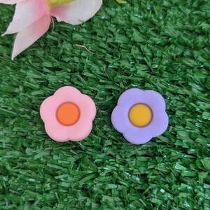 May include: Two small flower-shaped buttons, one pink with an orange center and one purple with a yellow center. The buttons are on a green surface.