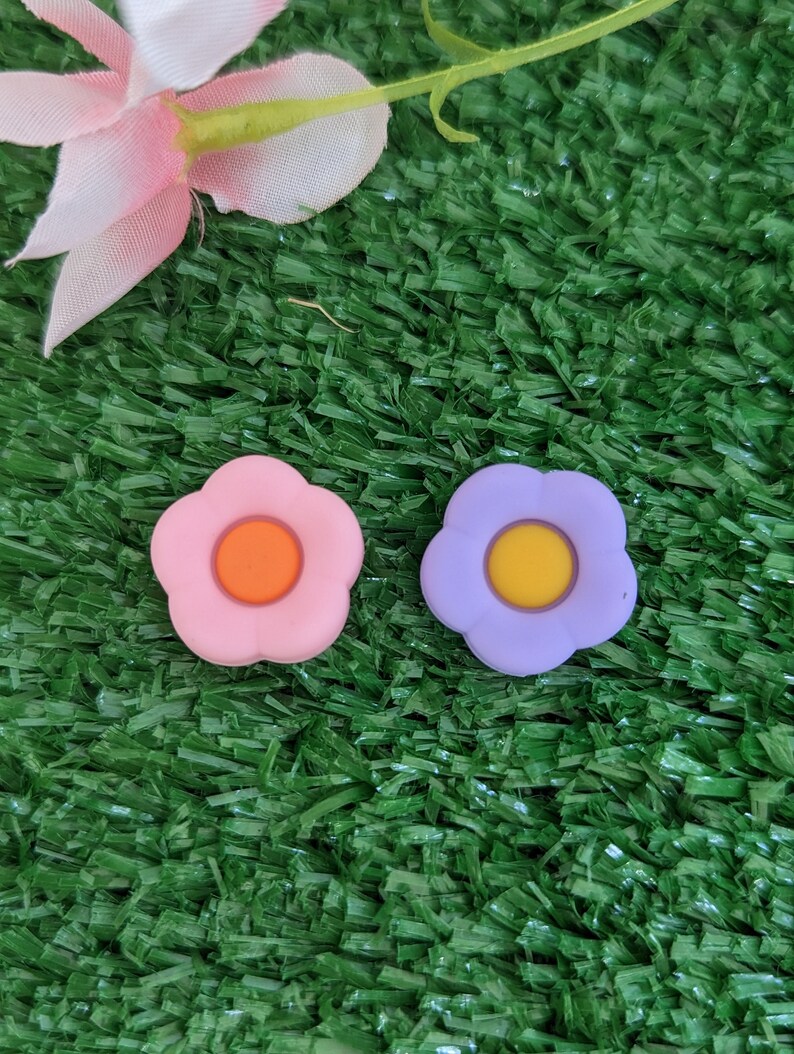 May include: Two small flower-shaped buttons, one pink with an orange center and one purple with a yellow center. The buttons are on a green surface.