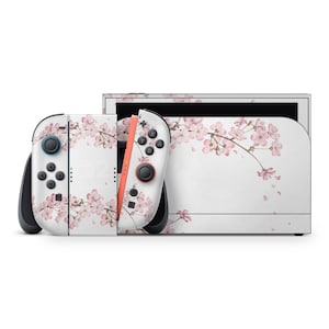 Op de afbeelding: Een witte gameconsole en controllers zijn versierd met een roze kersenbloesemdesign. De controllers hebben zwarte knoppen en joysticks, met een koraalkleurige rail. De console heeft een zwart scherm.