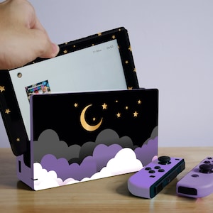 Pode incluir: Um console Nintendo Switch roxo e preto com um design de céu noturno estrelado. O console está aberto e a tela mostra um fundo branco. Também são mostrados dois controladores Joy-Con roxos e pretos.