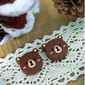 Pu&ograve; includere: Due decorazioni a forma di orso marrone con guance rosa e naso e bocca beige, appoggiate su una tovaglietta di pizzo bianco. Sullo sfondo una pigna e un oggetto sfocato marrone e bianco.