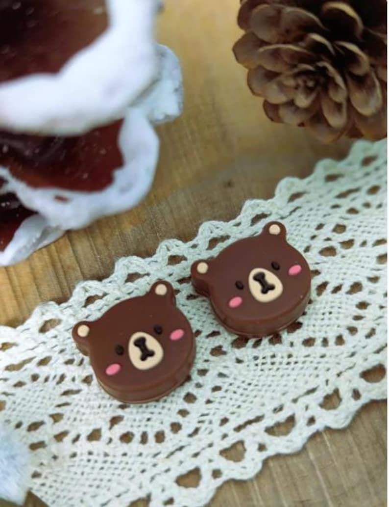 Pu&ograve; includere: Due decorazioni a forma di orso marrone con guance rosa e naso e bocca beige, appoggiate su una tovaglietta di pizzo bianco. Sullo sfondo una pigna e un oggetto sfocato marrone e bianco.