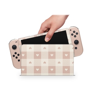 May include: A pink and white checkered Nintendo Switch case with a pink and white heart pattern. The case is open and a hand is holding the Nintendo Switch console.