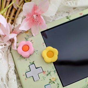 Può includere: Una console Nintendo Switch Lite verde e floreale con un pulsante a forma di fiore rosa e arancione e un pulsante a forma di fiore giallo.