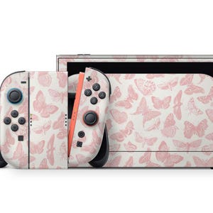 May include: A Nintendo Switch console and controllers featuring a pink butterfly design on a white backdrop. The controllers have black buttons and joysticks. The console has a black screen.