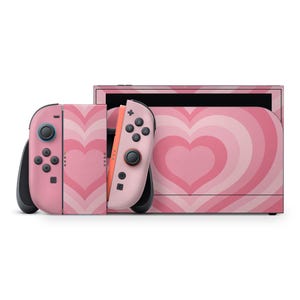 May include: A pink video game console with matching controllers. The console and controllers feature a repeating heart pattern in shades of pink and white. The controllers have black accents and buttons.