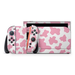 May include: A Nintendo Switch console and Joy-Con controllers with a pink and white cow print design. The controllers have black buttons and joysticks. The console is white with a black screen.