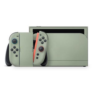 Könnte beinhalten: Eine salbeigrüne Spielekonsole mit zwei abnehmbaren Controllern. Der linke Controller hat blaue Tasten, während der rechte Controller rote Tasten hat. Die Konsole ist für den Handheld- oder Docking-Betrieb konzipiert.