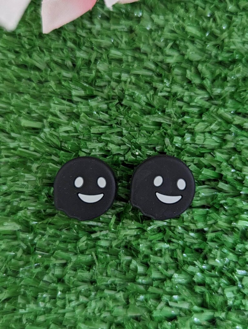 May include: Two black ghost-shaped silicone charms with white smiling faces. The charms are on a green artificial grass background.