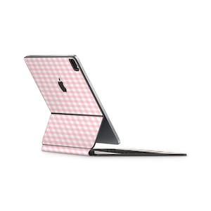 Pode incluir: Uma capa para iPad com padrão vichy rosa e branco com um teclado correspondente. A capa está fechada e o teclado está preso à parte de trás do iPad.