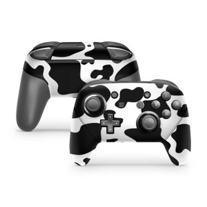 Op de afbeelding: Een zwart-witte Nintendo Switch-controller met een koe-print. De controller heeft een zwart-witte koe-print met zwarte knoppen en een zwarte joystick.