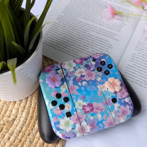 Puede incluir: Un mando Nintendo Switch Joy-Con azul y morado con un diseño floral y tapas de botones rosas. El mando está sobre una superficie tejida.