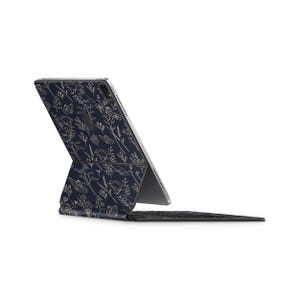 Puede incluir: Una funda de tableta azul marino con un estampado floral en beige claro. La funda está abierta, revelando una tableta y un teclado. El diseño floral incluye varias flores y hojas. El teclado es negro.