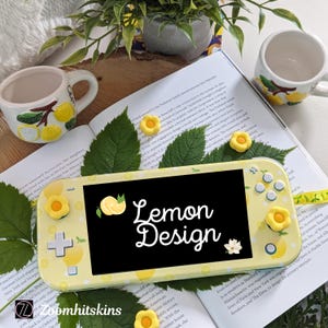 May include: A yellow and white Nintendo Switch Lite console with a lemon design. The console has a screen that says "Lemon Design" and is decorated with lemon slices and flowers. The console is on a book with green leaves.