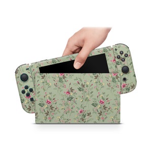 May include: A green Nintendo Switch console with a floral pattern. The console is open and the screen is visible. The console is being held by a hand.