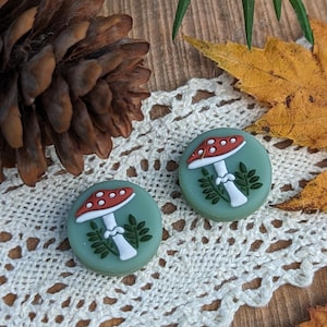 Op de afbeelding: Twee ronde oorbellen met paddenstoelmotieven. Elke oorbel heeft een rood-witte paddenstoelkap, een witte steel en groene bladeren. Ze liggen op een wit kanten kleedje, met een houten achtergrond en herfstbladeren.