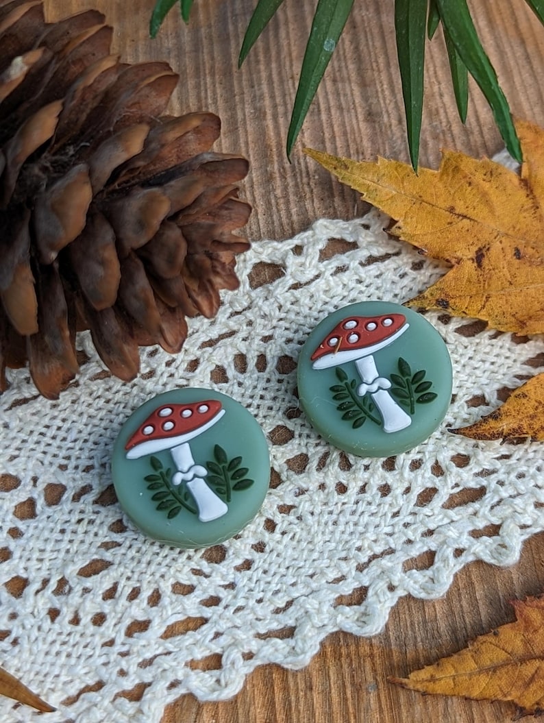 Op de afbeelding: Twee ronde oorbellen met paddenstoelmotieven. Elke oorbel heeft een rood-witte paddenstoelkap, een witte steel en groene bladeren. Ze liggen op een wit kanten kleedje, met een houten achtergrond en herfstbladeren.
