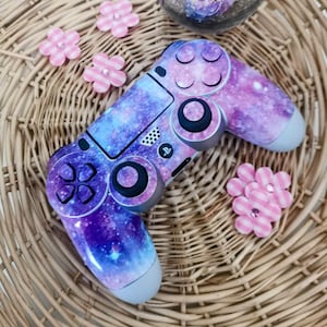 Puede incluir: Un mando de PlayStation 4 con temática de galaxia en colores morado, azul y rosa. El mando tiene una superficie texturizada y está decorado con un estampado de galaxia.