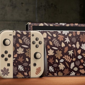 May include: A beige Nintendo Switch console and controller with a brown and beige leaf pattern. The console and controller are on a wooden surface.