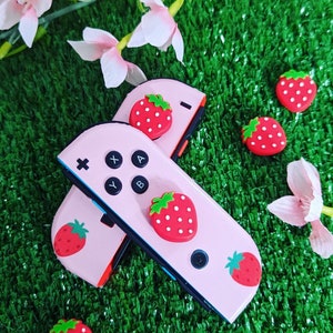 Może przedstawiać: Różowe kontrolery Joy-Con do Nintendo Switch z nakładkami na przyciski w kształcie truskawek i wzorem truskawki z tyłu. Kontrolery Joy-Con leżą na zielonej powierzchni z różowymi kwiatami.