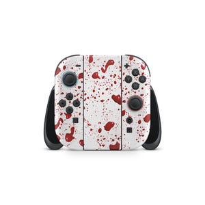 May include: A white Nintendo Switch controller with a pattern of red splatters. The controller has black joysticks and buttons, with a red button on the right side. The design covers the entire surface of the controller.