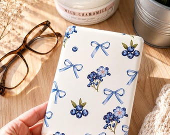 Può includere: Una custodia rettangolare bianca per e-reader con un motivo ripetuto di fiocchi azzurri, grappoli di mirtilli blu scuro con foglie verdi e piccoli fiori blu. Il design è stampato sulla superficie liscia della custodia protettiva.