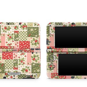 Puede incluir: Una consola de juegos portátil con una piel de patchwork con un patrón de fresas, flores y cuadros vichy en tonos rojo, verde, rosa y crema. El dispositivo se muestra tanto en la vista exterior cerrada como en la vista interior abierta con pantallas.