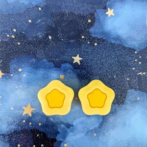 May include: Two yellow star-shaped hair clips against a blue and white watercolor background with gold glitter stars.