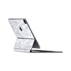 Può includere: Un iPad Pro Apple con una custodia dal design floreale in bianco e azzurro. La custodia è collegata a una tastiera nera. Il logo Apple è visibile sul retro dell'iPad.