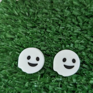 May include: Two white ghost-shaped charms with black eyes and a smiling mouth, set against a green artificial grass background.