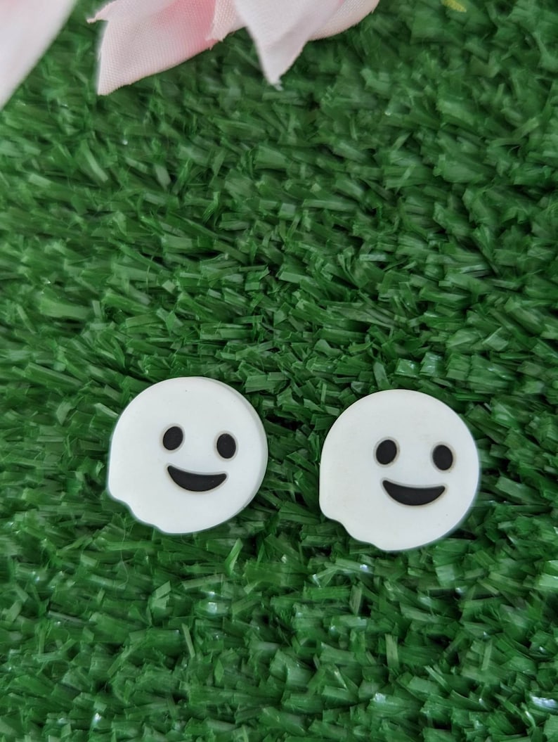 May include: Two white ghost-shaped charms with black eyes and a smiling mouth, set against a green artificial grass background.