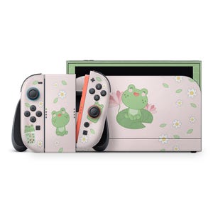May include: A Nintendo Switch console and controllers with a light pink frog-themed design. The design features cartoon frogs, flowers, and leaves. The controllers have black and orange accents.