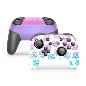 May include: Two wireless video game controllers. One controller is solid lavender with a pink top panel and black grips. The second controller features a white faceplate covered in a repeating pattern of purple and cyan leaf icons.
