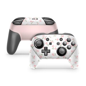 Op de afbeelding: Twee draadloze gamecontrollers. Eén is lichtroze met zwarte accenten. De andere is wit met een bloemmotief van roze rozen en groene bladeren. Beide controllers hebben zwarte knoppen en joysticks.