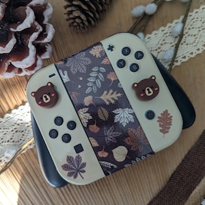 Può includere: Un controller Nintendo Switch beige con un design a foglie autunnali marroni e bianche. Il controller ha due pulsanti a forma di orso marrone.