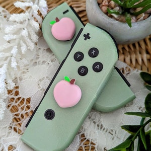 May include: A pair of green Nintendo Switch Joy-Con controllers with pink peach-shaped button covers. The controllers are on a white lace surface with a potted plant in the background.