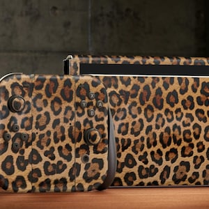 May include: A video game console and a handheld controller featuring a brown, black, and tan leopard print skin. The controller has black joysticks and buttons, while the console dock displays the same animal pattern across its front surface.