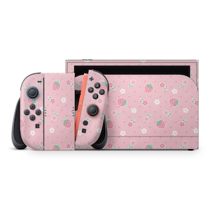 May include: A pink Nintendo Switch console and controllers with a strawberry and flower pattern. The controllers are detached, and the console is in the dock. The design features a repeating pattern of strawberries and white flowers.