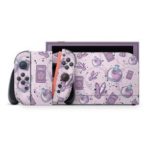 Könnte beinhalten: Eine hellviolette Spielkonsole mit passenden Controllern. Konsole und Controller sind mit einem Muster aus Zaubertränken, Kristallen und Tarotkarten verziert. Die Controller haben schwarze Knöpfe und eine korallenfarbene Schiene.