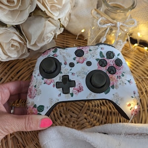 以下が含まれることがあります： ピンクのバラと緑の葉が描かれた花柄の白いビデオゲームコントローラー。コントローラーは黒いボタンとトリガーが付いています。
