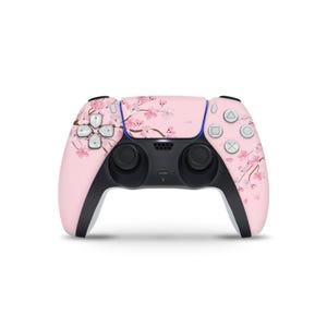 May include: A pink PlayStation 5 controller with a floral cherry blossom design. The controller has black handles and buttons, with a blue accent. The face of the controller features a pattern of pink flowers and branches.