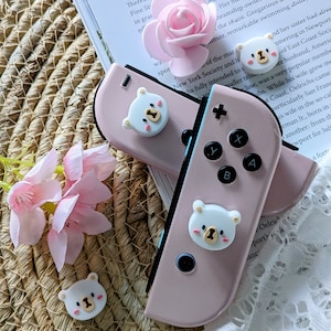 Op de afbeelding: Een roze en blauwe Nintendo Switch-console met witte beer-vormige joy-con-covers. De console ligt op een geweven strooppervlak met roze bloemen.