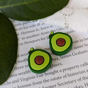 May include: Two green avocado-shaped charms with brown pits. The charms are made of a soft, pliable material and are perfect for adding a touch of whimsy to your crafts.